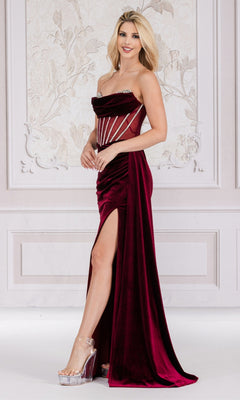 A woman in the Sheer-Waist Strapless Long Velvet Prom Dress 5051—featuring a sheer beaded corset bodice and high slit—stands before an ornate, light-colored wall, finishing her elegant look with clear high heels.