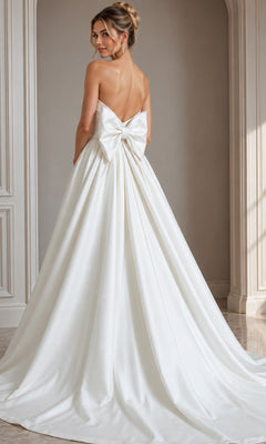 A woman in a strapless satin gown with a dramatic back bow stands in an elegant room, facing away from the camera—a graceful showcase of designer formal dress style, embodied by the Long Formal Dress: Jovani 50534.