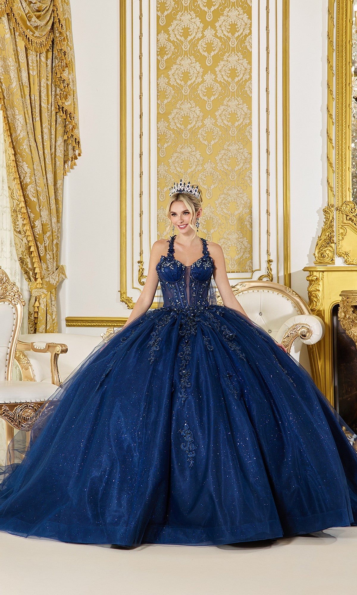 A woman in the Quinceanera Dress 5061 by Dancing Queen, a navy gown with beaded applique and a tiara, stands in an ornate gold-decorated room featuring a chair, drapes, and a fireplace.
