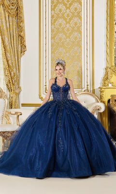 A woman in the Quinceanera Dress 5061 by Dancing Queen, a navy gown with beaded applique and a tiara, stands in an ornate gold-decorated room featuring a chair, drapes, and a fireplace.