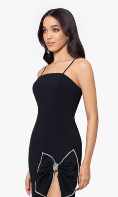 Woman wearing the Long Prom Dress 5063BN by Blondie Nites, a fitted black formal dress with thin straps and rhinestone bow detail at the hip, standing against a plain white background.
