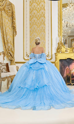 A woman in the Quinceanera Dress 5064 by Dancing Queen, featuring a light blue sheer corset bodice, sits facing away in an ornate room with gold accents, a fireplace, and a chandelier.