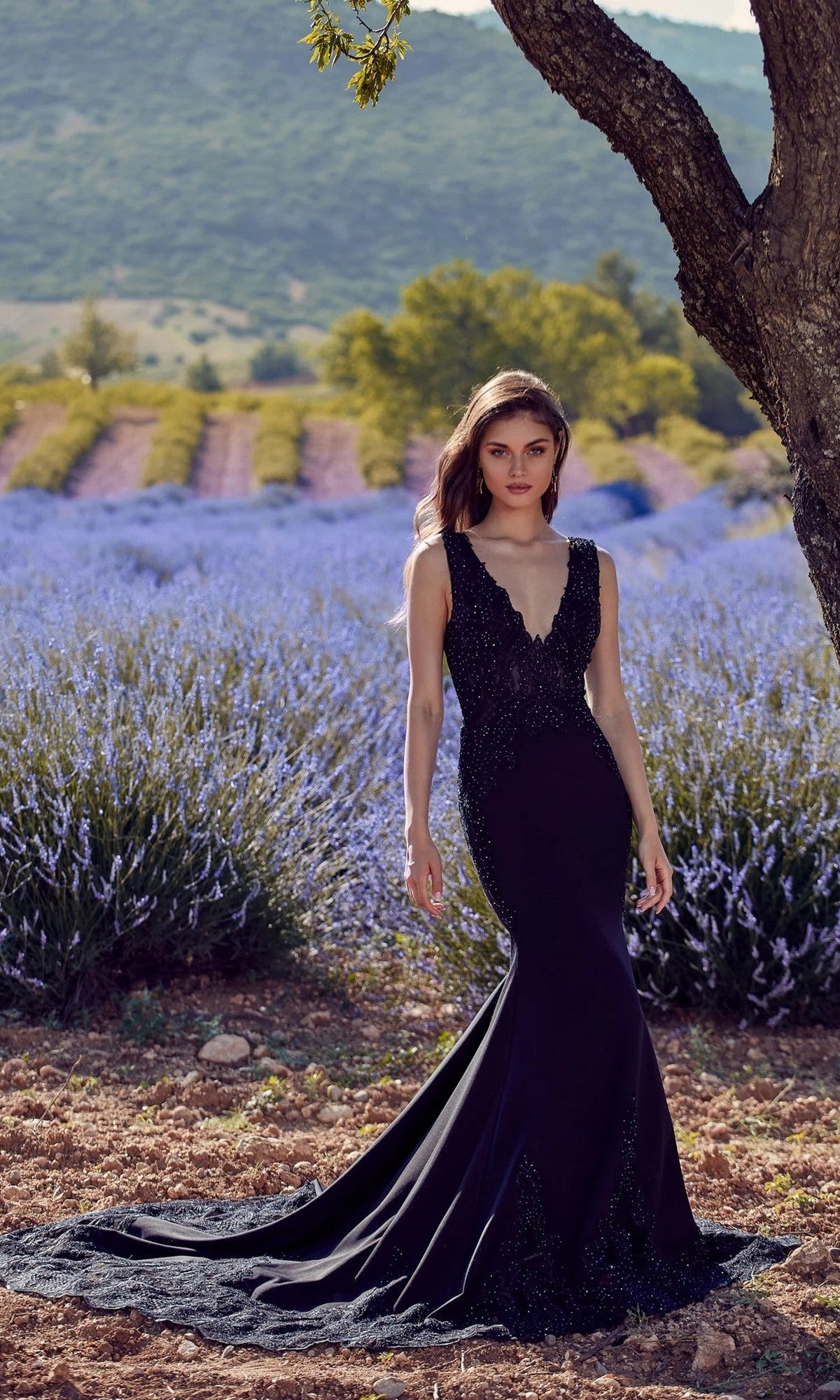 Wearing the Alyce 5065 long formal dress in black, a woman stands by a tree outdoors, with lavender fields and distant hills behind her. The sleeveless gown features a fitted silhouette and lace-embellished bodice.