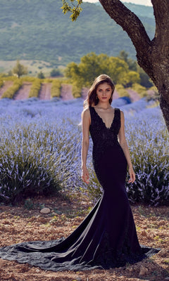 Wearing the Alyce 5065 long formal dress in black, a woman stands by a tree outdoors, with lavender fields and distant hills behind her. The sleeveless gown features a fitted silhouette and lace-embellished bodice.