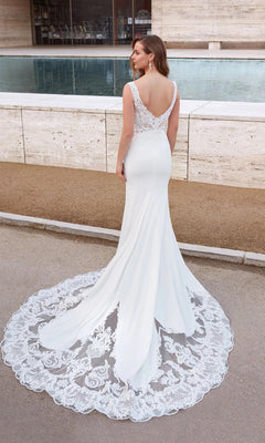A woman poses by a pool in the Alyce 5065 long formal dress featuring a fitted white silhouette, lace-embellished bodice, open back, and elegant train.