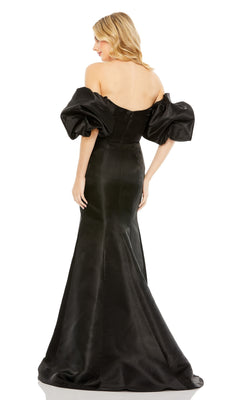 A woman stands facing away in the Off-the-Shoulder Long Gala Gown, Mac Duggal 50677, showcasing its fitted back, voluminous short sleeves, and flared skirt that evoke classic mermaid evening gown elegance.