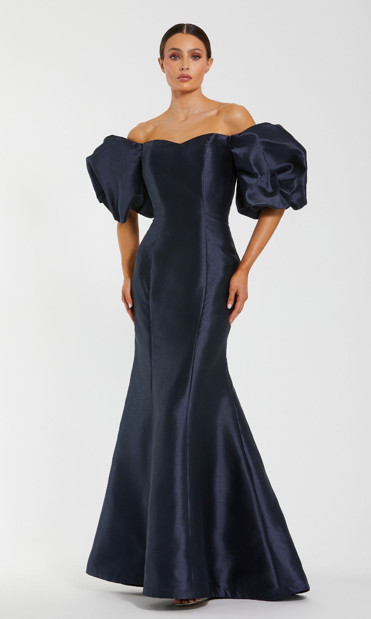 A woman wears the Mac Duggal 50677 Off-the-Shoulder Long Gala Gown in dark navy, featuring a fitted mermaid silhouette with dramatic puff sleeves and a flared hem, standing against a plain light background.