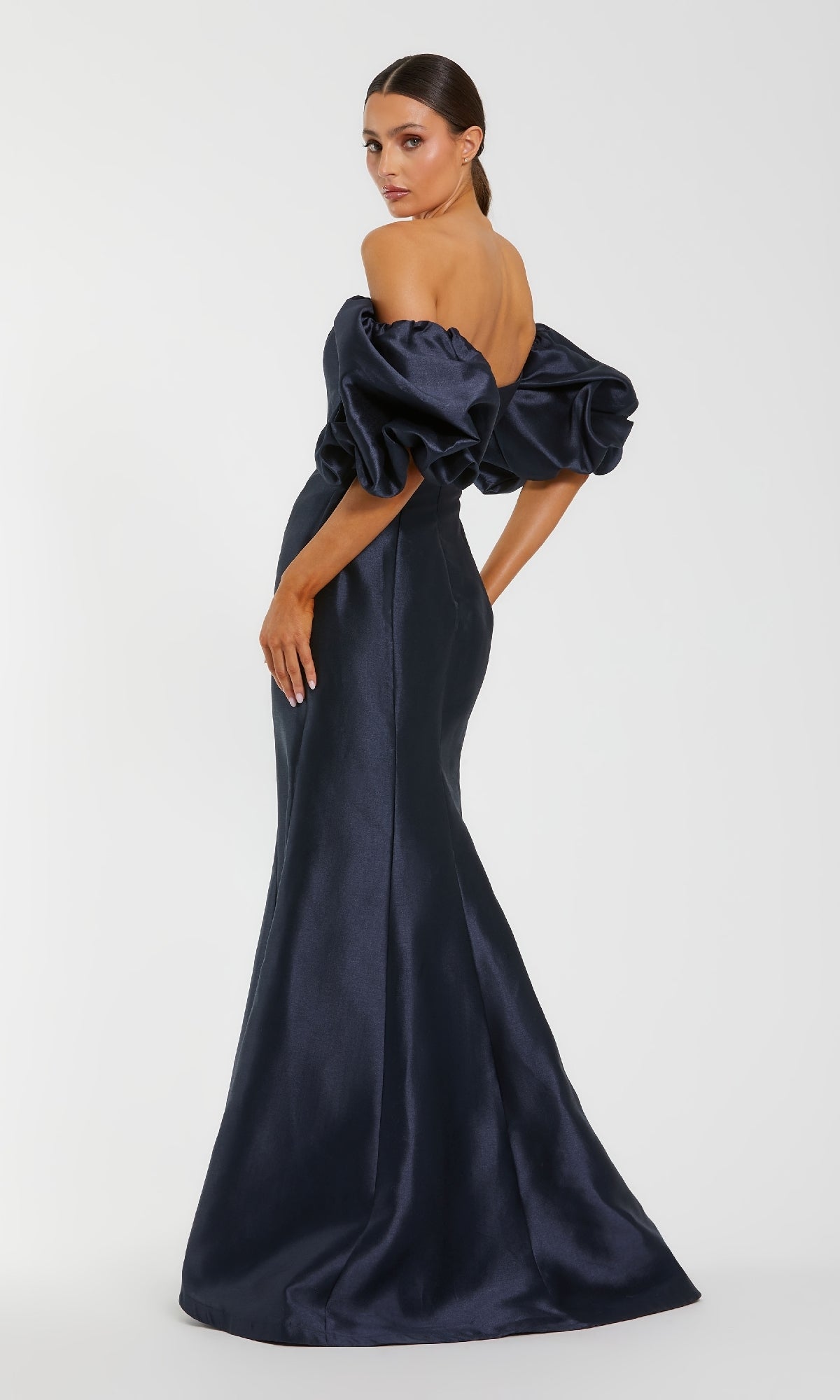 A woman poses against a plain white background wearing the Mac Duggal 50677 Off-the-Shoulder Long Gala Gown—a dark blue, floor-length formal dress with puffed sleeves.