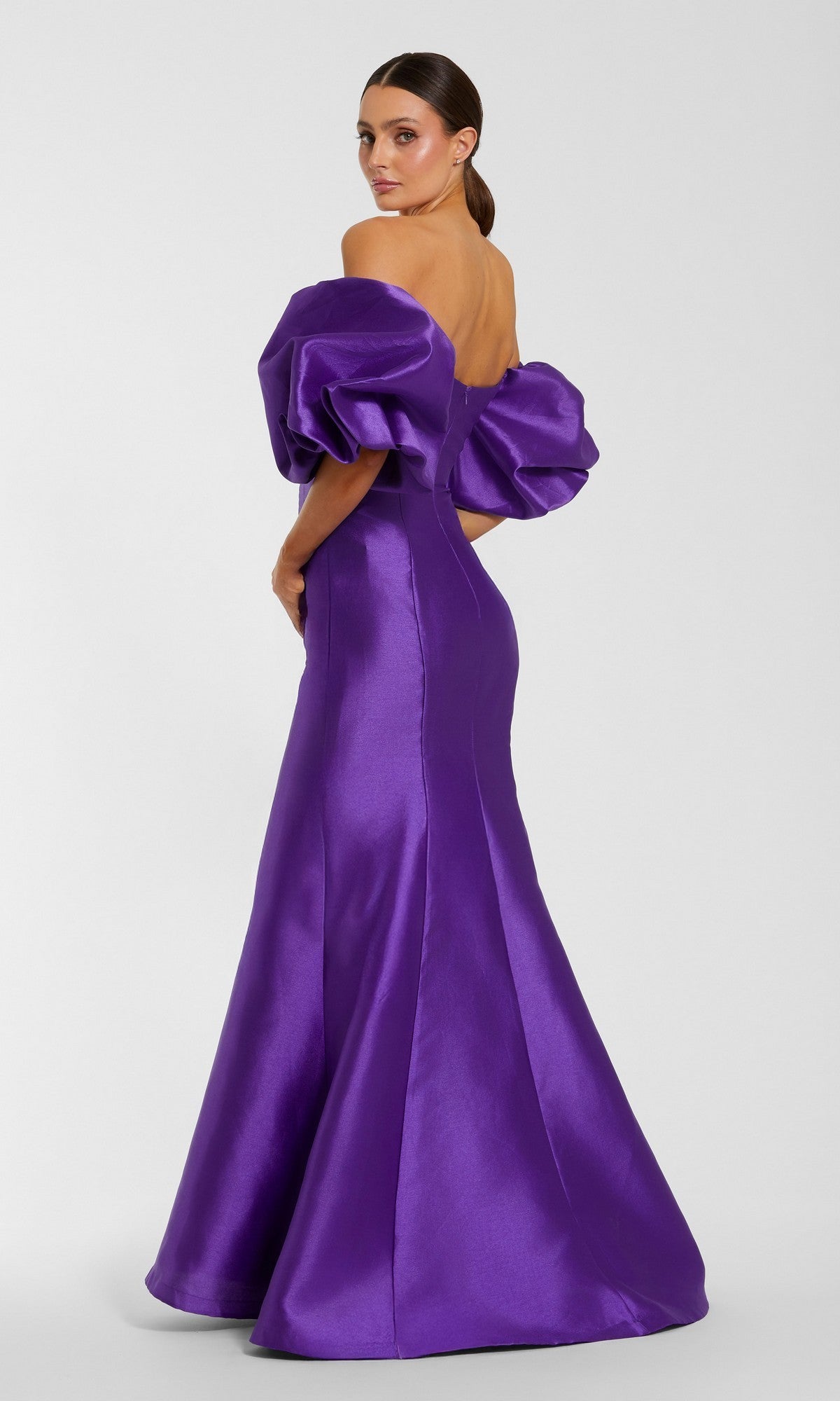 A woman stands against a plain background, looking over her shoulder in the Mac Duggal 50677 Off-the-Shoulder Long Gala Gown, featuring dramatic puff sleeves and a shiny purple mermaid silhouette.