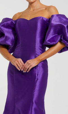 A woman stands with hands clasped, wearing the Mac Duggal 50677 Off-the-Shoulder Long Gala Gown—a fitted purple satin mermaid dress featuring voluminous puff sleeves.
