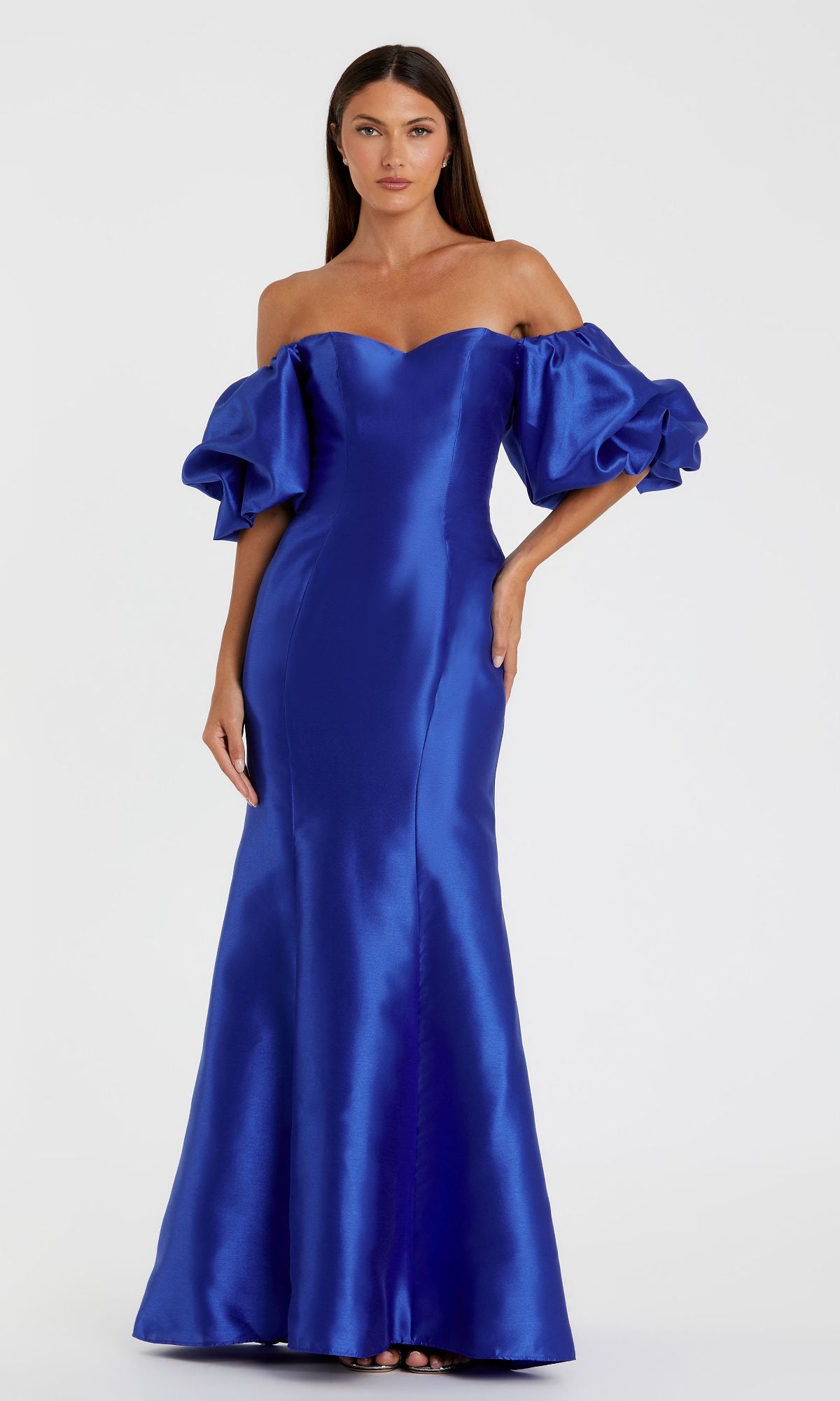 Woman in the Mac Duggal 50677 off-the-shoulder long gala gown, featuring a shiny royal blue mermaid silhouette with puffed sleeves, stands against a plain light background.