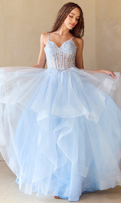 A woman stands indoors against a beige wall, wearing the Amelia Couture 5068 Long Glitter Prom Ball Gown—a light blue, layered tulle dress with a fitted, embellished bodice and spaghetti straps.