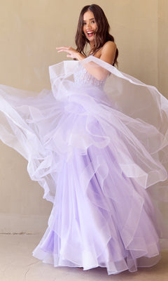 A woman twirls in the Long Glitter Prom Ball Gown: Amelia Couture 5068, her light purple a-line dress sparkling as its layers flare out and she smiles against a plain beige wall.