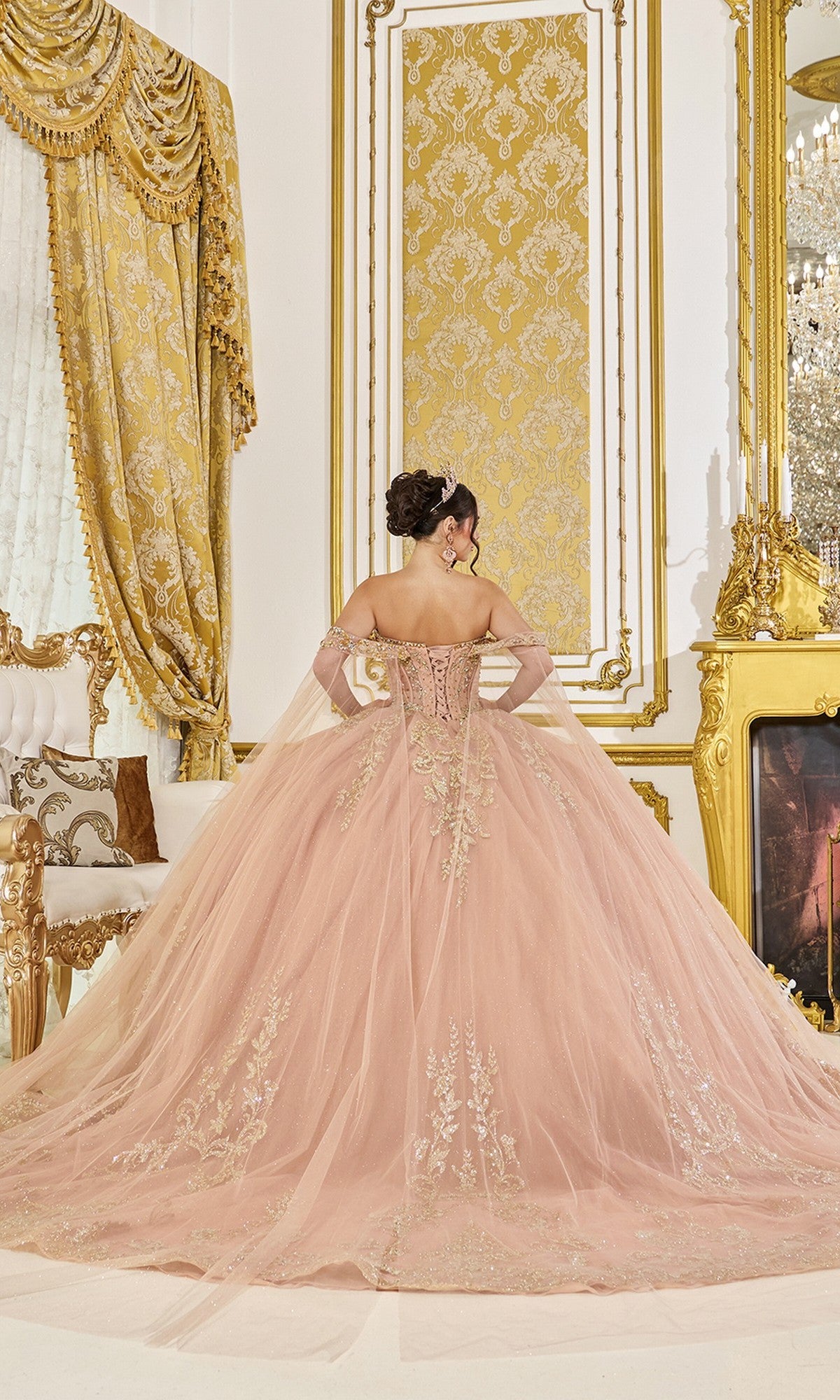 A woman in the Quinceanera Dress 5069 by Dancing Queen—a pink off-the-shoulder ballgown with gold embroidery and glitter tulle—stands in a lavish, gold-decorated room, facing away from the camera.