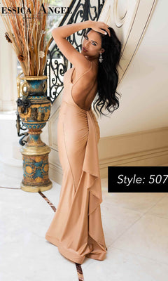A woman in a backless beige gown poses indoors beside an ornate vase and railing. Style: Jessica Angel 507. The fitted Back-Sash Long Prom Dress radiates elegance—ideal for those desiring the distinctive allure of Jessica Angel designs.