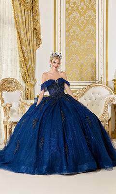 A woman wearing the navy Quinceanera Dress 5075 by Dancing Queen, featuring glitter tulle and off-the-shoulder sleeves, poses with a tiara in an ornate gold and white room with an elaborate chair, curtain, and wall paneling.