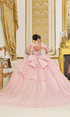 A woman wears the Quinceanera Dress 5079 By Dancing Queen, featuring blush pink fabric and a large bow, as she stands with her back to the camera in an ornate room with gold accents.