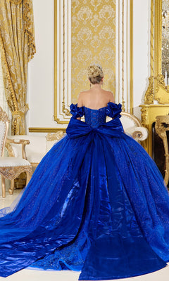 A woman with blonde hair in an updo models the Quinceanera Dress 5080 By Dancing Queen, a royal blue off-the-shoulder gown with a large bow, standing in an ornate gold and white room, seen from behind.
