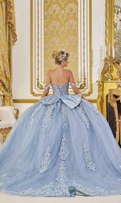 A woman wears the Quinceanera Dress 5087 By Dancing Queen—a light blue gown with lace details and a large bow—standing in an ornate gold and white room, facing away from the camera.