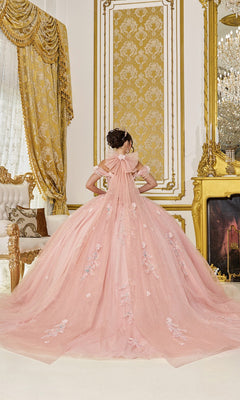 A person in the Quinceanera Dress 5096 By Dancing Queen, featuring blush pink glitter tulle and floral embellishments, stands before an ornate gold-trimmed wall in a lavishly decorated room.