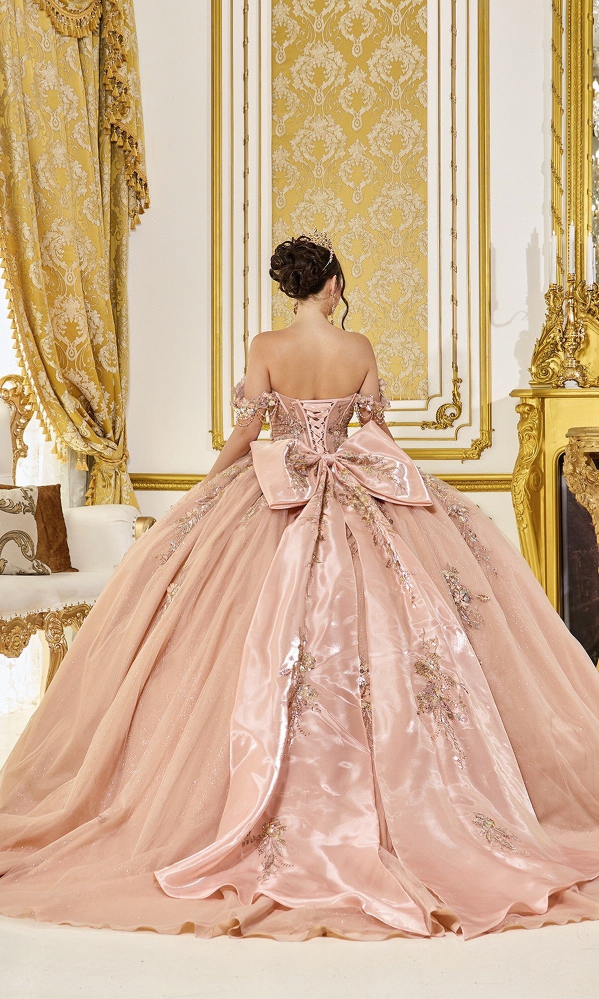 A woman wearing the Quinceanera Dress 5097 by Dancing Queen—a pink off-the-shoulder gown adorned with floral embroidery, an embellished corset bodice, and a dramatic back bow—stands in an ornate room accented with gold decor.