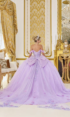 A woman in the Quinceanera Dress 5098 by Dancing Queen, featuring 3D floral accents and a sheer corset bodice, stands in an ornate gold-decorated room with her back to the camera.