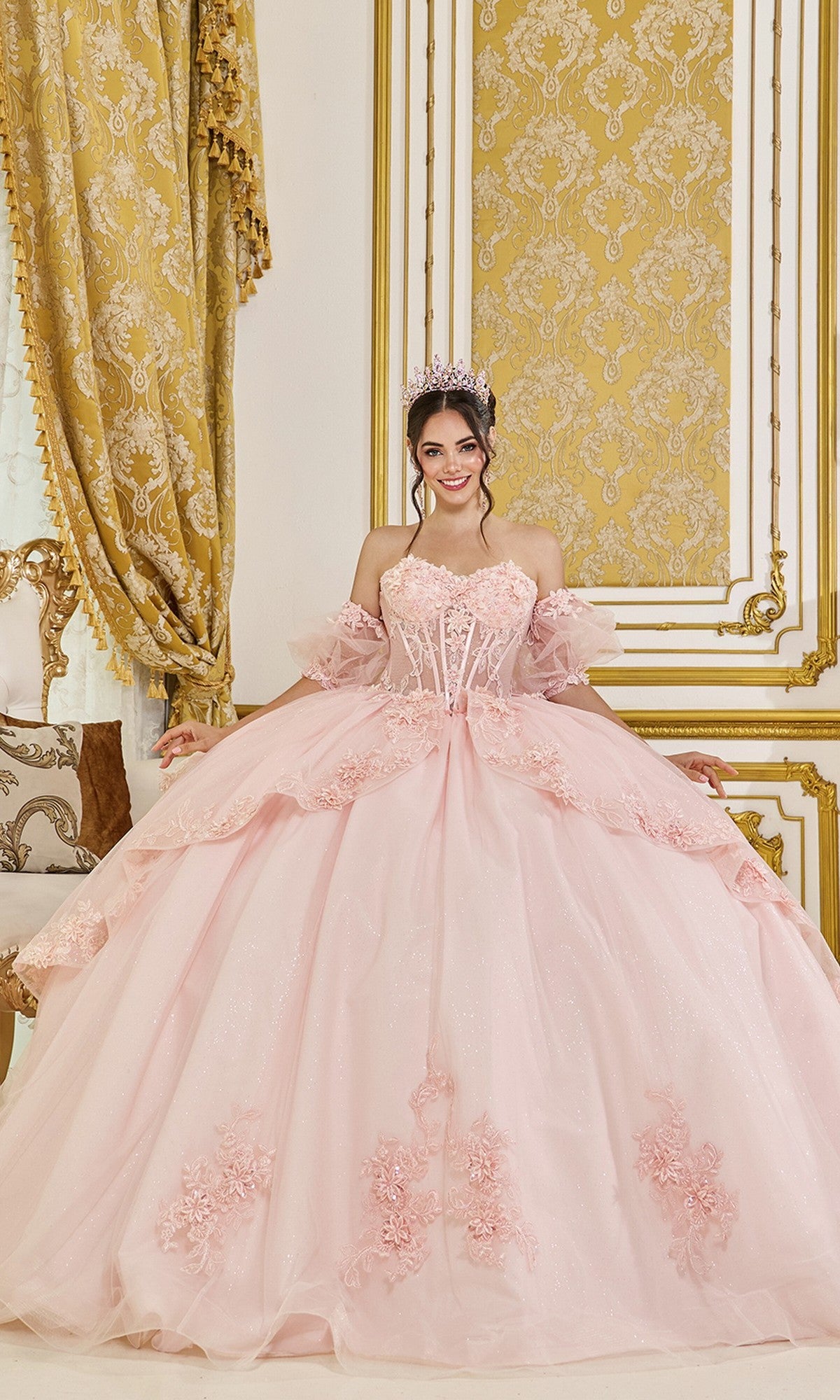 A woman in the Quinceanera Dress 5099 by Dancing Queen in blush, adorned with floral details and a tiara, stands in an ornately decorated room with gold and cream accents.