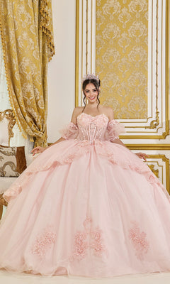 A woman in the Quinceanera Dress 5099 by Dancing Queen in blush, adorned with floral details and a tiara, stands in an ornately decorated room with gold and cream accents.