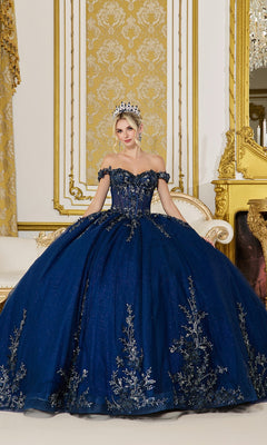 A woman poses in an ornate gold room wearing the Quinceanera Dress 5102 By Dancing Queen in navy, featuring silver lace embroidery and a tiara.