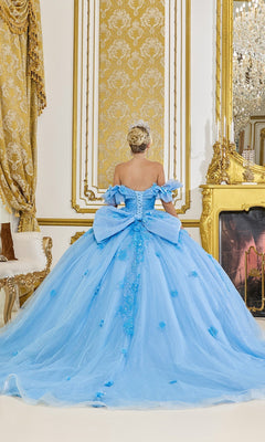 A person wearing the Quinceanera Dress 5103 By Dancing Queen, a stunning blue gown with off-the-shoulder sleeves and a large bow, stands facing away in an ornate, gold-accented room. The dress features a voluminous ballgown tulle skirt that adds ethereal elegance to the scene.