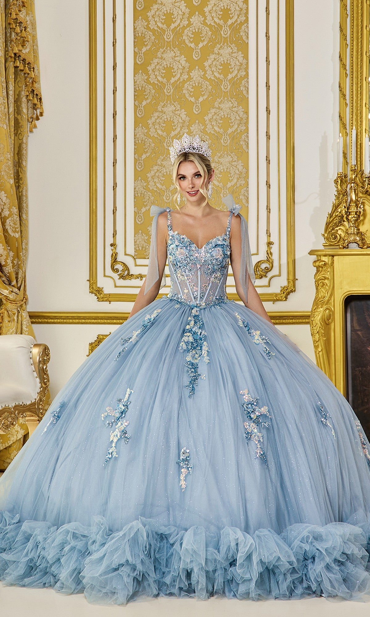 A woman in the Quinceanera Dress 5104 by Dancing Queen, a dusty blue glitter tulle ball gown embellished with 3D flowers, stands in an ornate gold and white room, wearing a jeweled tiara.