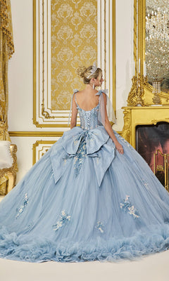 A woman stands in an ornate gold and white room, facing away from the camera, wearing the Quinceanera Dress 5104 By Dancing Queen—a light blue gown adorned with 3D flowers and a large bow.