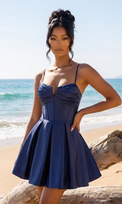 A woman in the Lace-Up Short Hoco Dress: Blondie Nites 5107BN in cadet blue stands by a driftwood log on a sandy beach, with the ocean and blue sky in the background.
