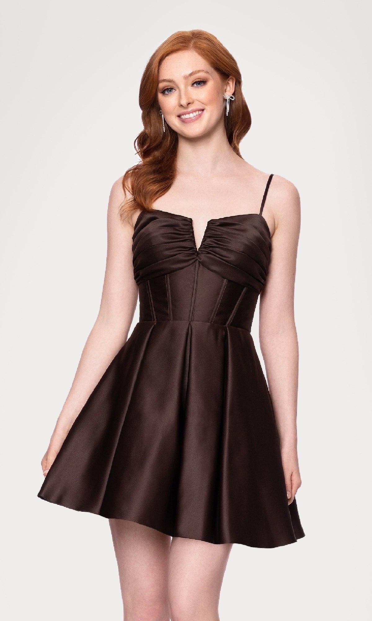 A woman wearing the Lace-Up Short Hoco Dress: Blondie Nites 5107BN—a sleeveless, dark brown fit-and-flare mini with thin straps and a lace-up back—stands and smiles against a plain background.