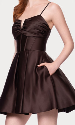 A woman wears the Lace-Up Short Hoco Dress: Blondie Nites 5107BN, a sleeveless dark brown satin mini with lace-up back, fitted bodice, thin straps, and flared skirt, posing with one hand in her pocket.