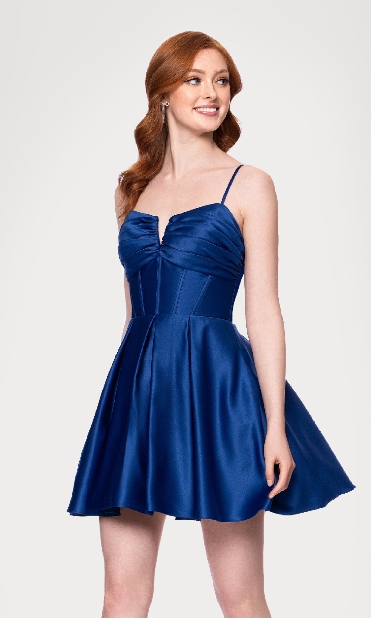 A woman with long red hair wears the affordable Blondie Nites 5107BN Lace-Up Short Hoco Dress—a sleeveless, fitted blue satin party dress with thin straps and a flared skirt—standing against a plain light background.