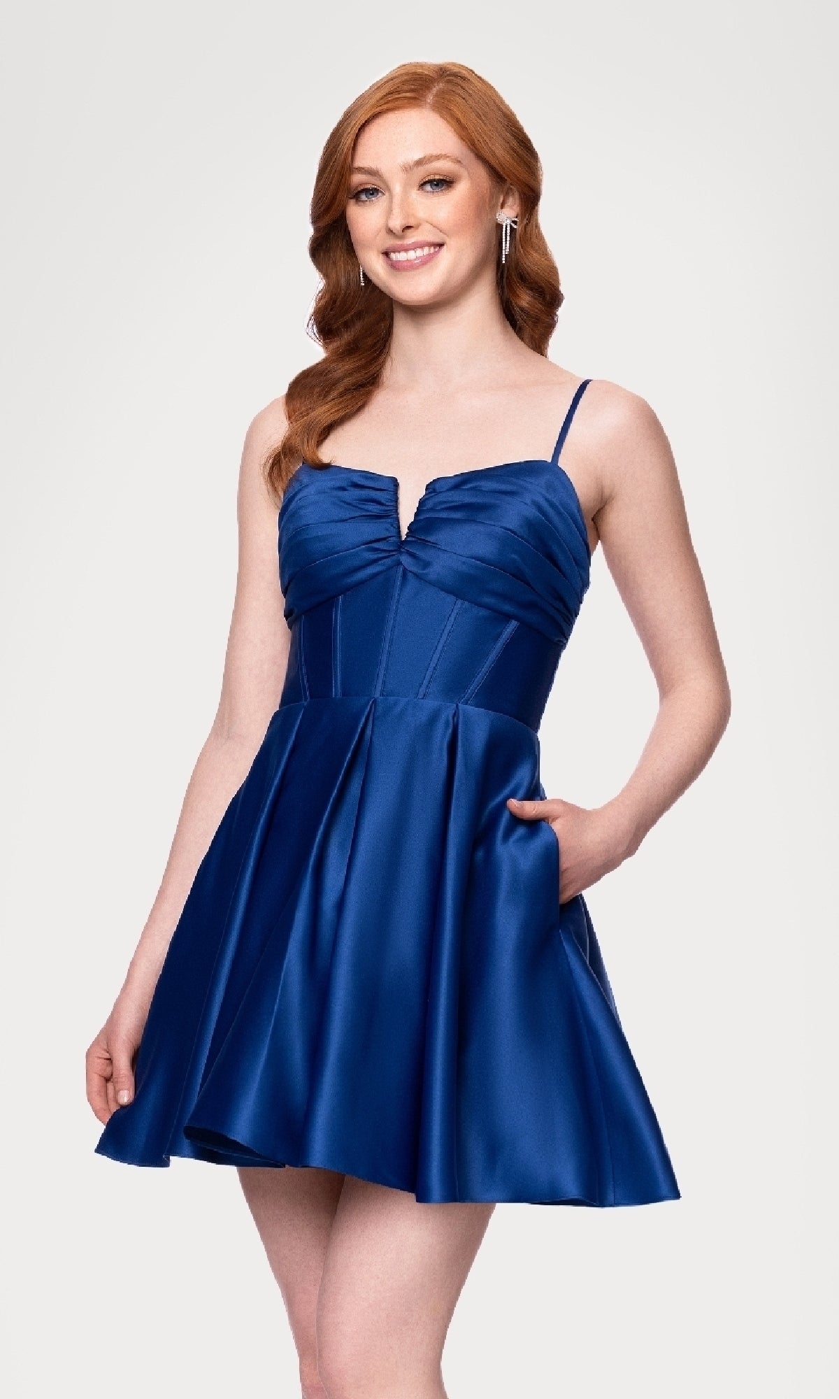 Woman wearing the Lace-Up Short Hoco Dress: Blondie Nites 5107BN—a sleeveless royal blue satin dress with spaghetti straps, V-neck, flared skirt, and lace-up back—smiling with one hand in her pocket.
