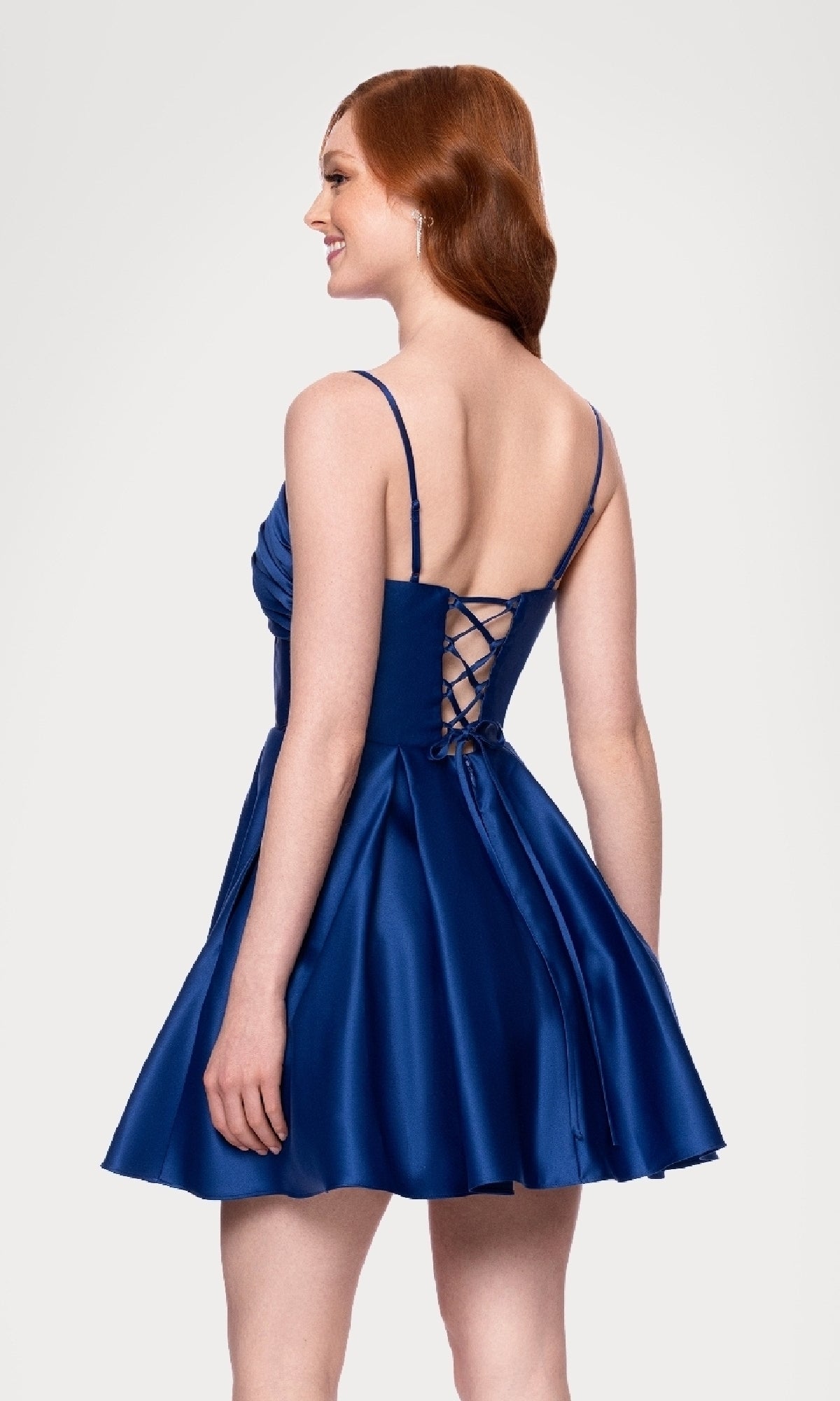 A woman in the affordable Blondie Nites 5107BN Lace-Up Short Hoco Dress—royal blue, sleeveless satin with a lace-up back and flared skirt—smiles as she stands, her head turned to the side.