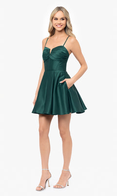 A woman stands against a plain white background wearing the Blondie Nites 5107BN Lace-Up Short Hoco Dress, an emerald green satin mini dress with thin straps, flared skirt, and lace-up back, paired with silver high-heeled sandals.