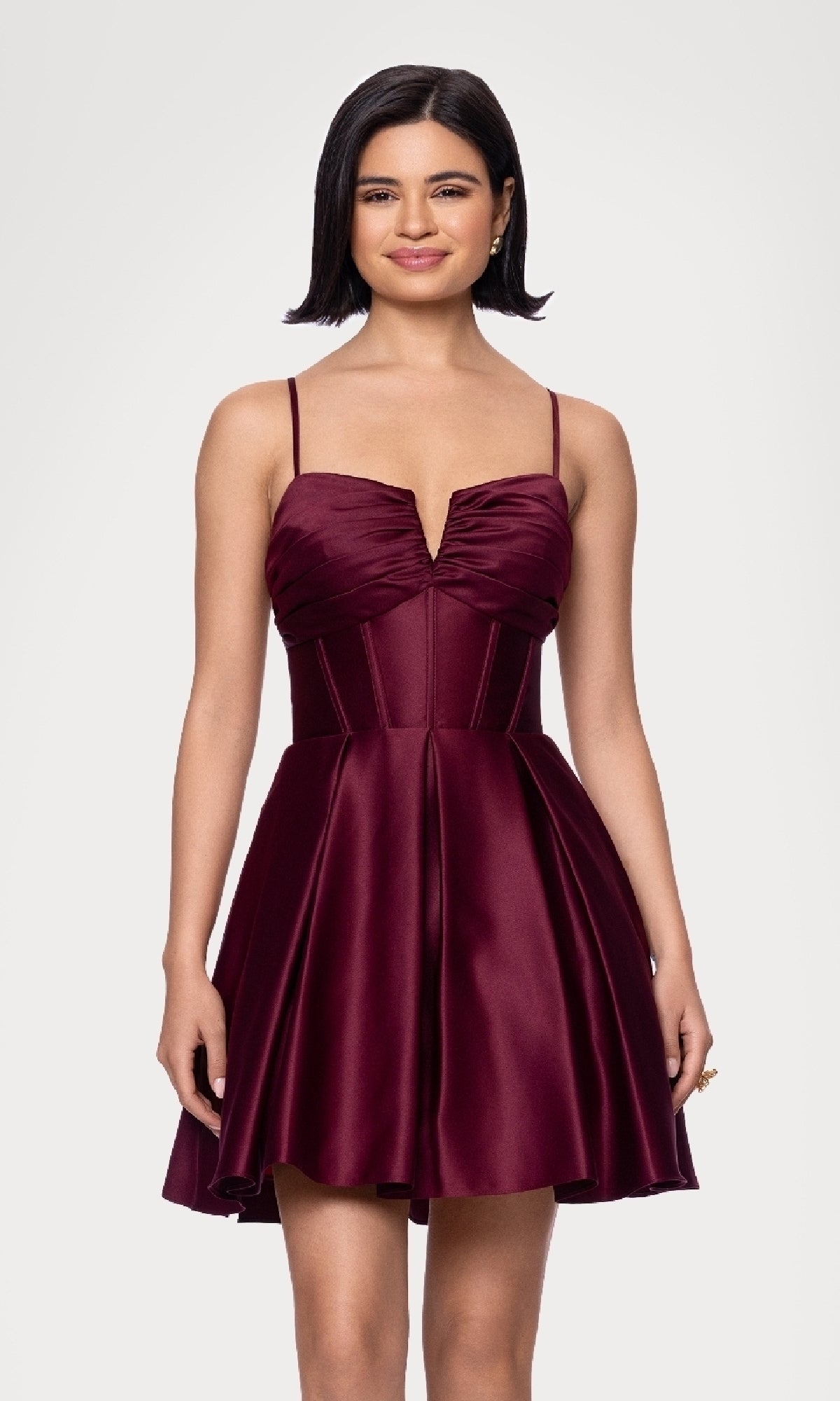 A woman with short dark hair models the Lace-Up Short Hoco Dress: Blondie Nites 5107BN, a sleeveless deep burgundy satin corset homecoming dress with a fitted bodice and flared skirt, standing against a plain light background.