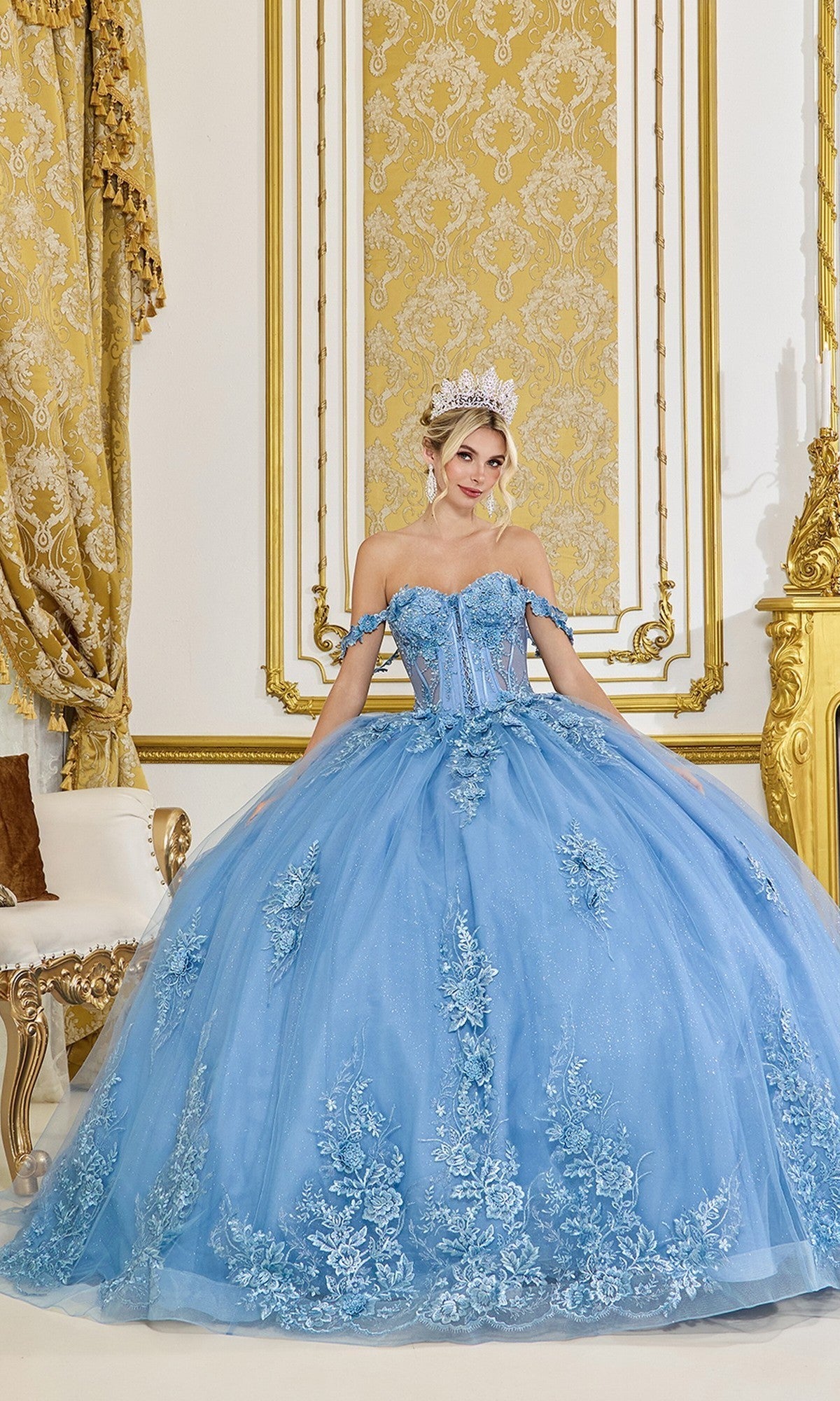 A woman stands in an opulent room adorned with gold decor and intricate curtains, wearing the Quinceanera Dress 5117 by Dancing Queen in Smoky Blue, featuring a glitter tulle ballgown skirt and an ornate corset bodice.