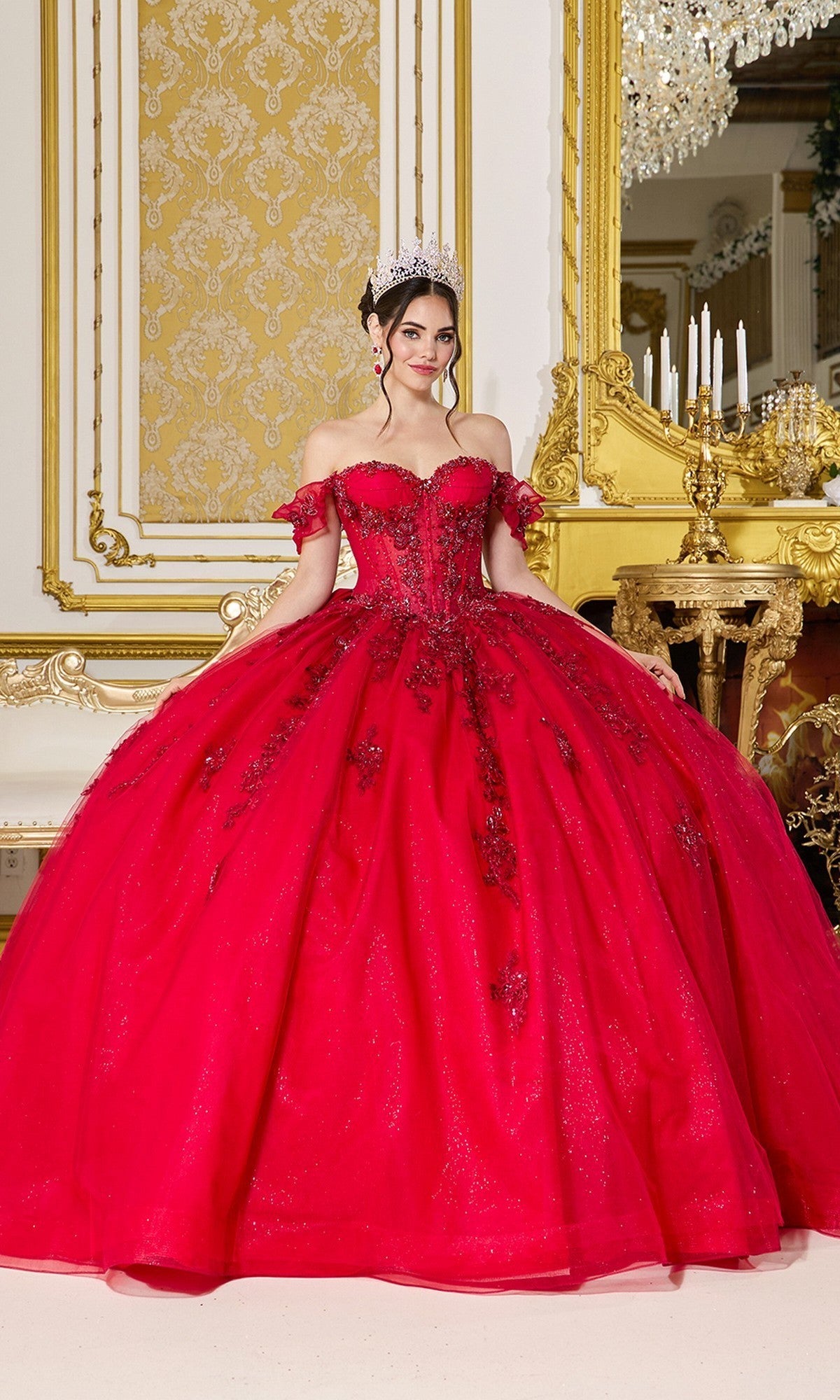 A woman is wearing the Quinceanera Dress 5118 by Dancing Queen in vibrant red, featuring a corset bodice and a tiara, as she stands in an opulent gold-trimmed room adorned with a candelabra and a large mirror behind her.