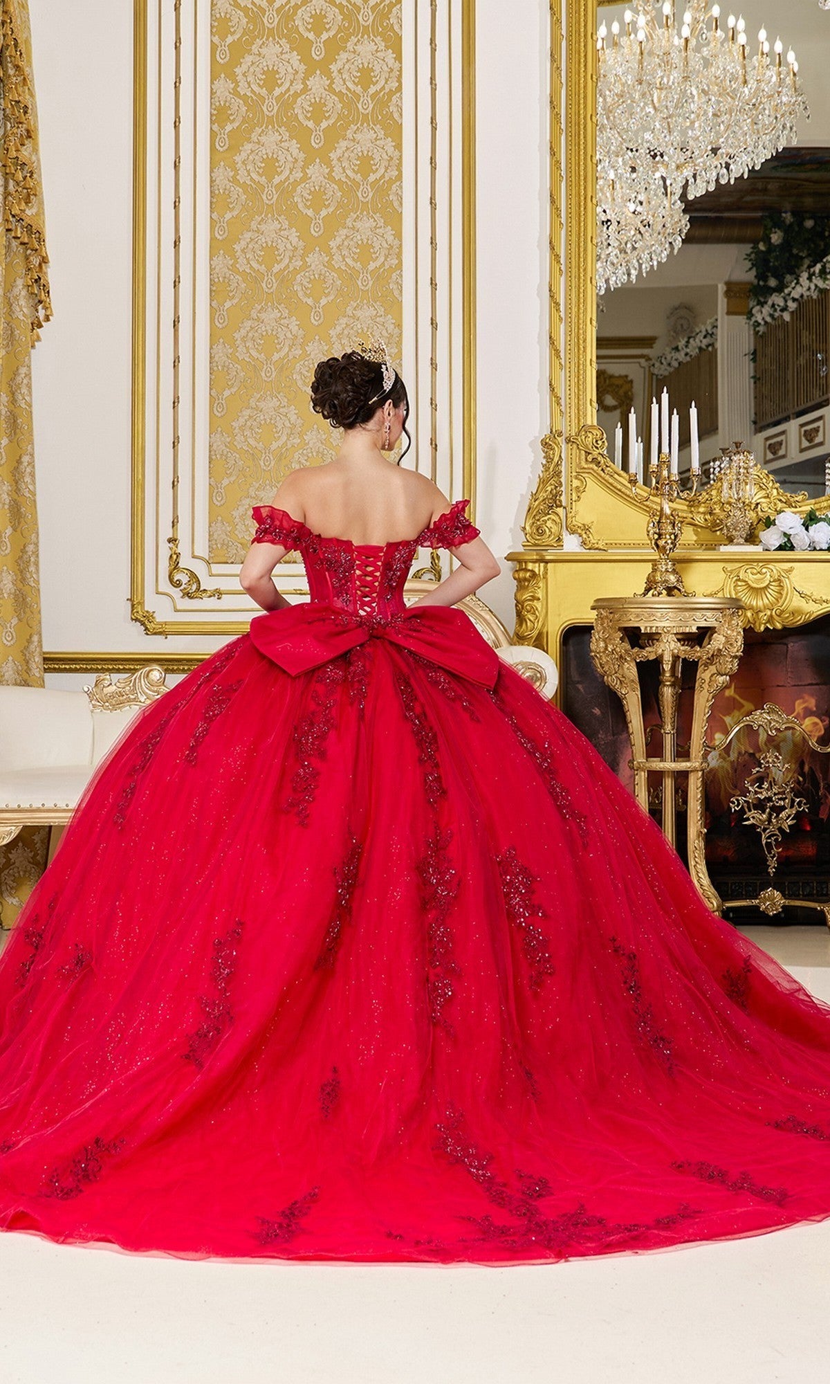 Wearing the Quinceanera Dress 5118 by Dancing Queen, a person stands facing a gold-framed mirror in an opulent room adorned with elaborate decor and a chandelier, showcasing the gown’s stunning glitter tulle, corset bodice, and dramatic large bow.