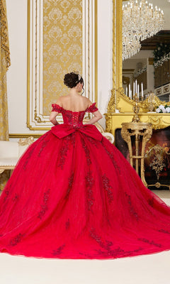 Wearing the Quinceanera Dress 5118 by Dancing Queen, a person stands facing a gold-framed mirror in an opulent room adorned with elaborate decor and a chandelier, showcasing the gown’s stunning glitter tulle, corset bodice, and dramatic large bow.