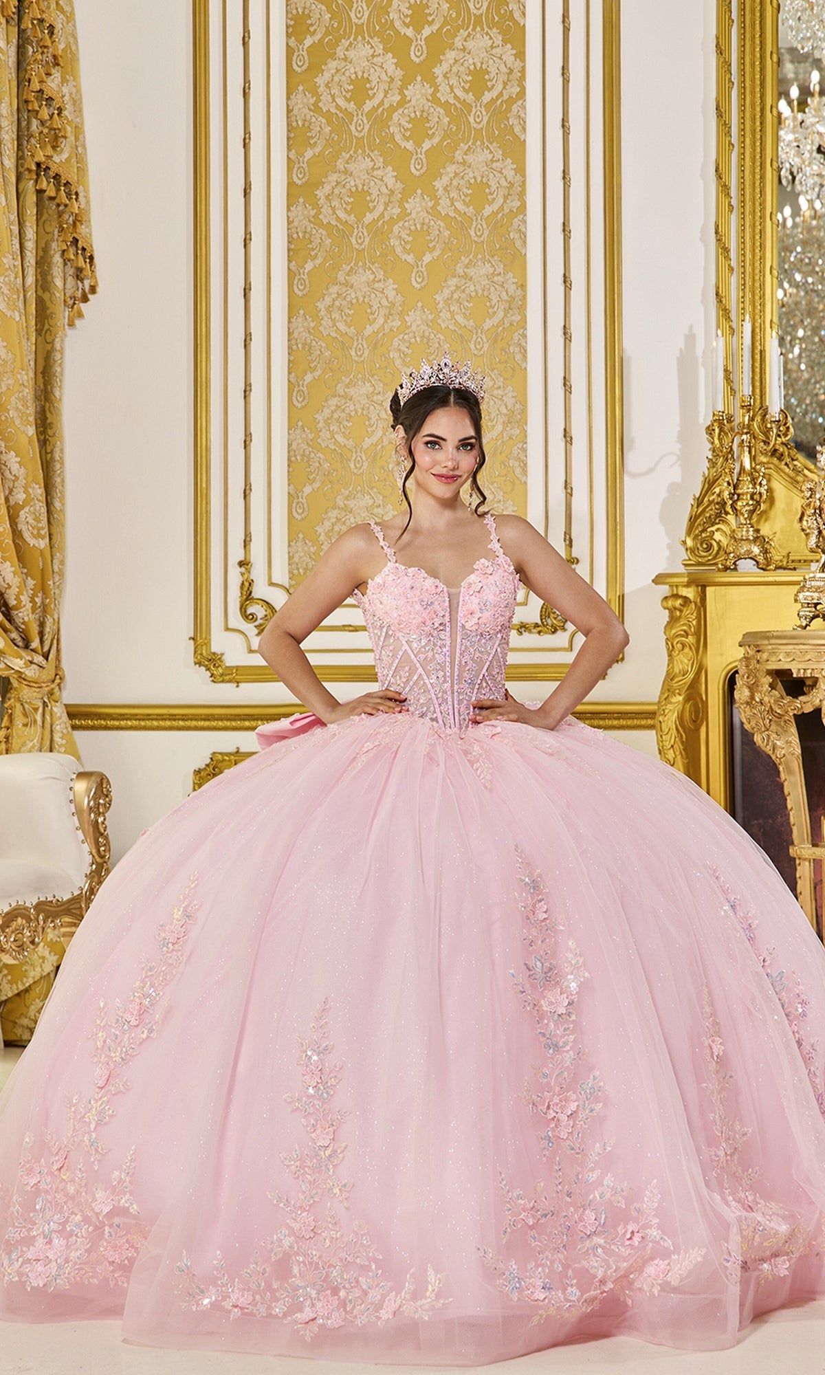A young woman stands smiling at the camera in an ornate, gold-decorated room, wearing the Quinceanera Dress 5119 By Dancing Queen in Blush. The pastel gown features delicate floral embellishments and a corset bodice, complemented by her sparkling tiara for a truly radiant look.
