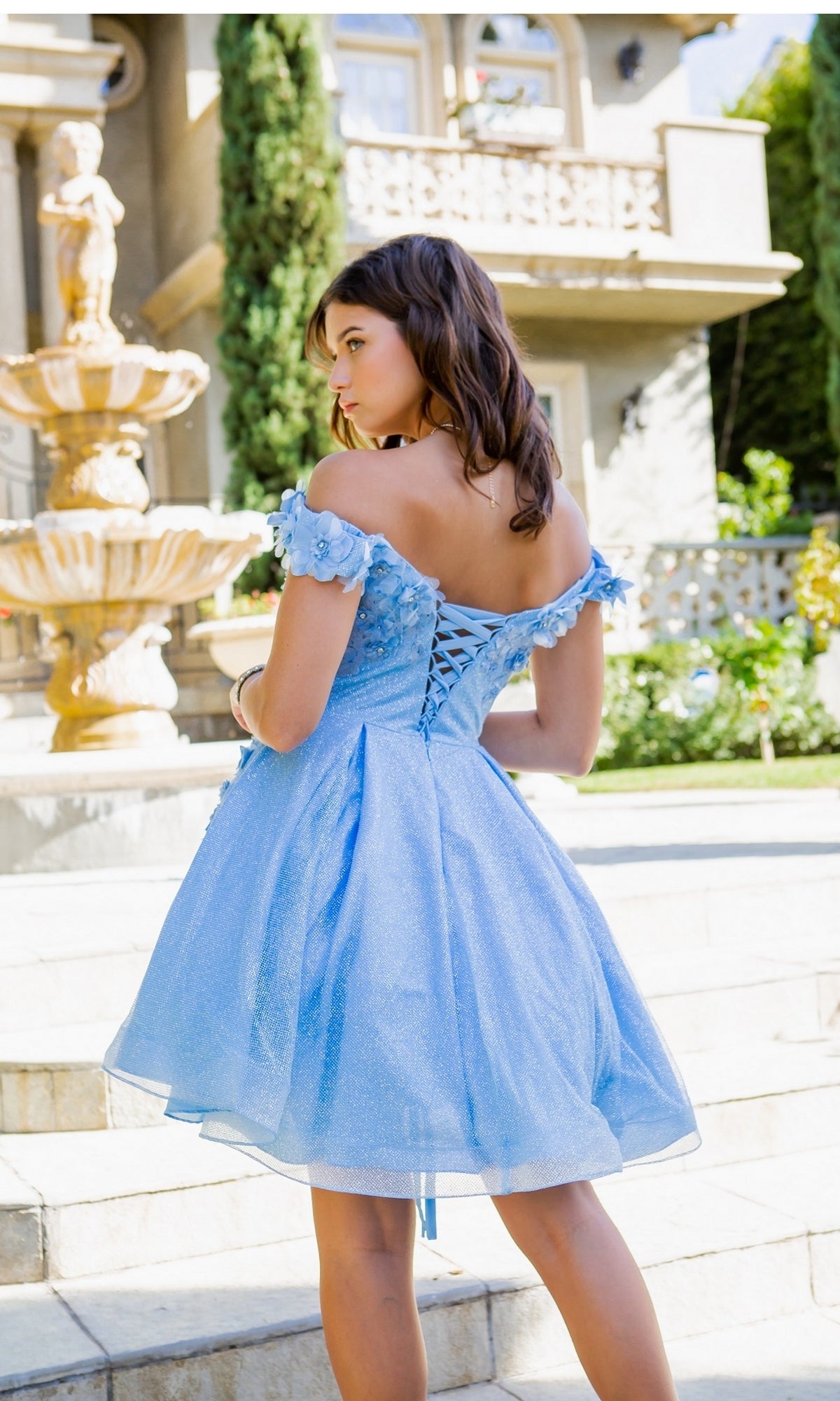 A young woman faces away from the camera outdoors, standing by a stone fountain and building, wearing the Shimmer Off-Shoulder Short Homecoming Dress 5120J in light blue.