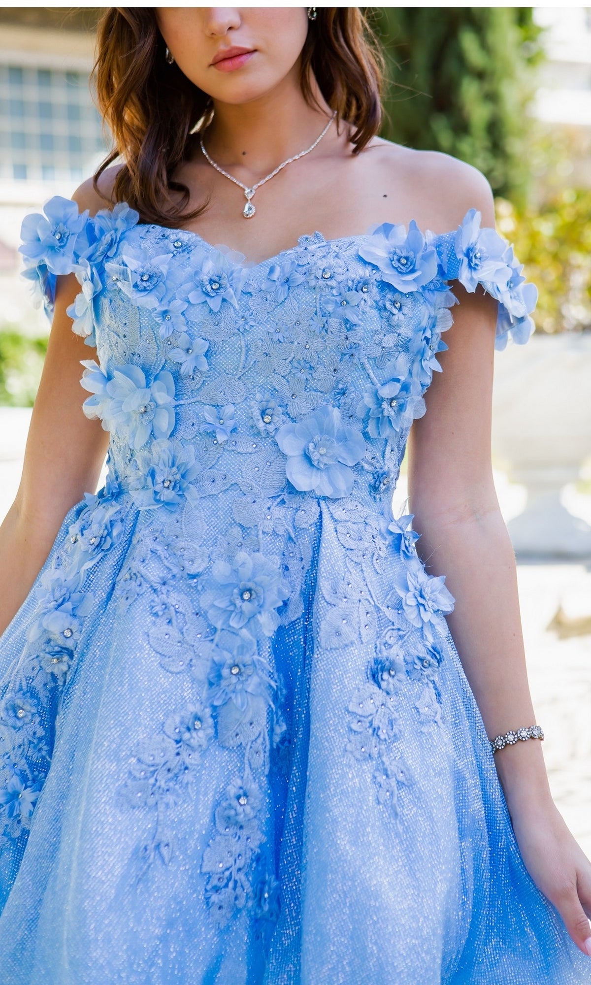 A woman wears the Shimmer Off-Shoulder Short Homecoming Dress 5120J in light blue, featuring 3D floral appliqués and lace, styled with a silver bracelet and necklace.