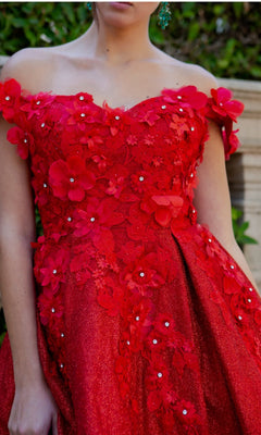 Wearing the Shimmer Off-Shoulder Short Homecoming Dress 5120J in red, featuring 3D floral appliqués and delicate embellishments, a person stands outdoors.