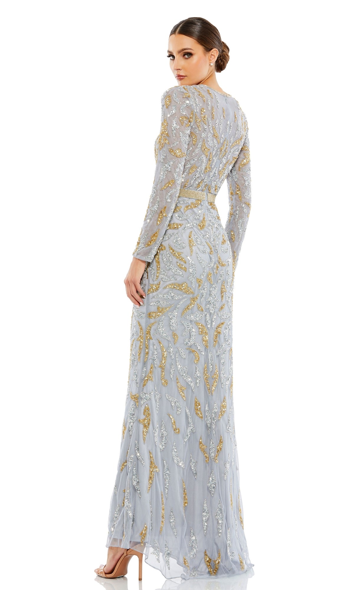 A woman stands looking over her shoulder, wearing the Mac Duggal 5124 Long Sleeve Sequin Formal Dress—floor-length with silver and gold sequins—styled elegantly with high-heeled sandals.