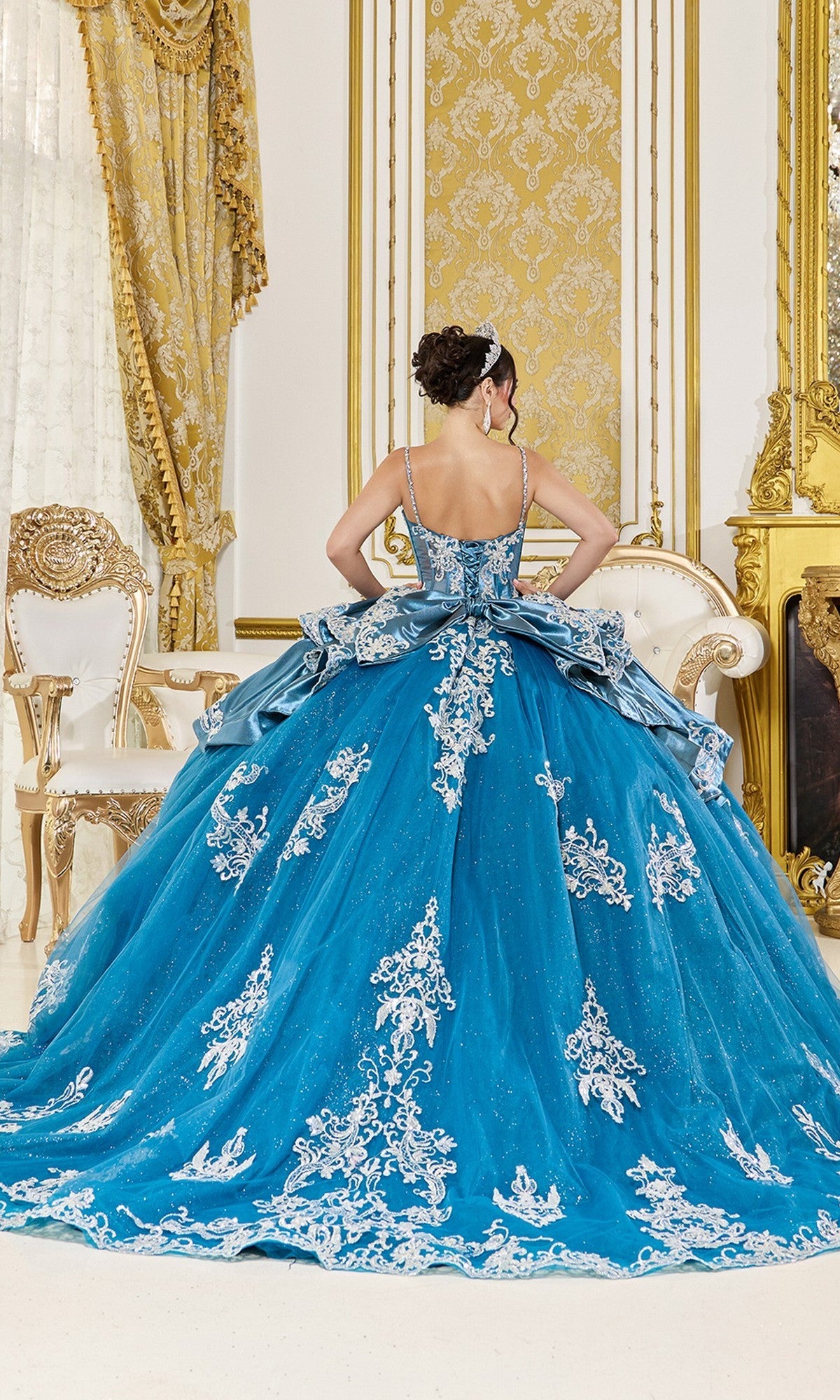 A woman in the Quinceanera Dress 5125 By Dancing Queen, featuring teal blue beaded applique, stands facing away in an ornate room with gold and white decor and vintage-inspired furniture.